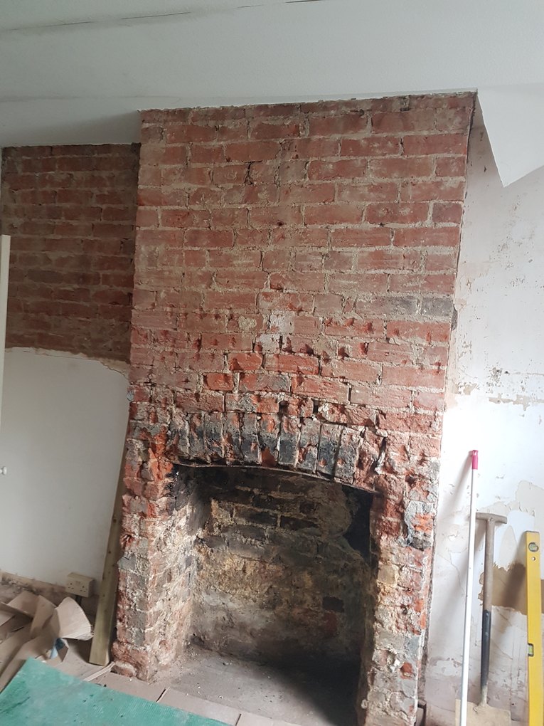 Exposed brick chimney breast and open fireplace before restoration, with plaster stripped back to bare brick